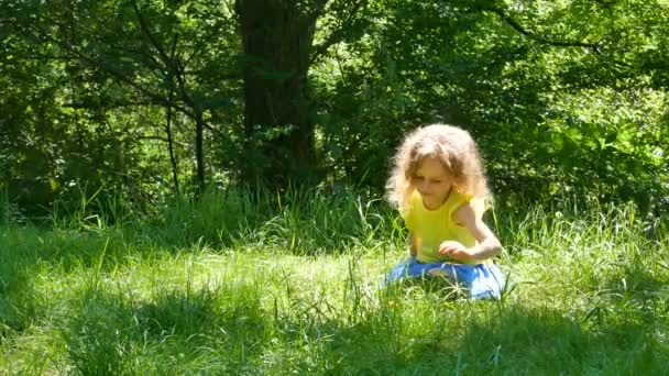 Retrato al aire libre de la hermosa niña con el pelo rizado corto rubio sentado en la hierba verde, recogiéndolo y vomitándolo . — Vídeo de stock