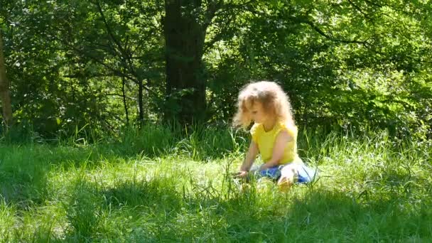 Petite fille ludique jouissant d'un temps chaud d'été En plein air dans le parc pendant la journée ensoleillée. Enfant femelle ramassant herbe verte et regardant la caméra .