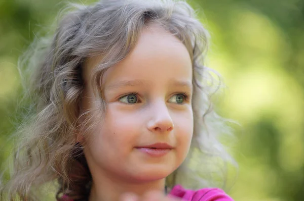 Belle petite fille blonde bouclée, a un visage souriant joyeux et joyeux, de grands yeux bleus, de longs cils. Portrait en bois vert — Photo