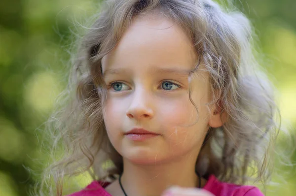 Belle petite fille blonde bouclée, a une expression sérieuse, regarde au loin, de grands yeux bleus, de longs cils. Portrait en fond de bois — Photo