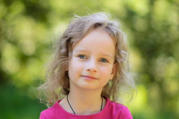 Gyönyörű göndör szőke kislány, már boldog szórakozás vidám mosolygós arc, nagy kék szemek, a hosszú szempillák. Portré-fa háttér — Stock Fotó