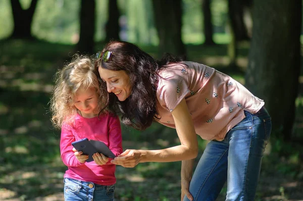 Joven morena madre está mostrando cómo utilizar el teléfono inteligente a su niño rubio disfrutando de un clima cálido y soleado fuera en el parque . — Foto de Stock