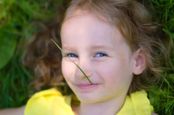 Küçük sarışın kız yalan yere ve Looking yeşil çimen kök ağız ile kamera var. Mavi gözlü kadın çocuk yüz closeup portresi. — Stok fotoğraf