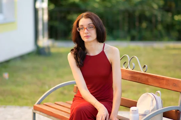 Молодая студентка в очках сидит на скамейке в солнечный день летом . — стоковое фото