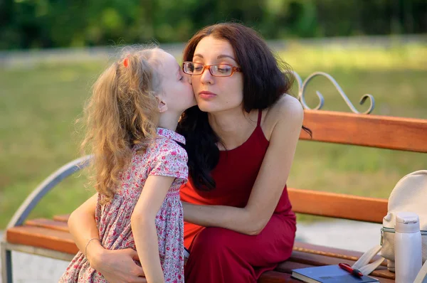 Cute Little Girl está besando a su madre sentada en el banco en el parque de la ciudad. Mujer joven con gafas de vista y vestido rojo. Niño tiene dos colas de caballo . —  Fotos de Stock