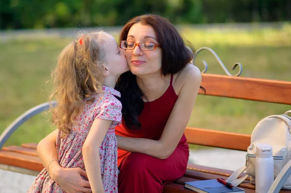 Mignonne petite fille blonde embrasse sa jeune mère brune en lunettes et robe rouge assise sur le banc dans le parc de la ville . — Photo