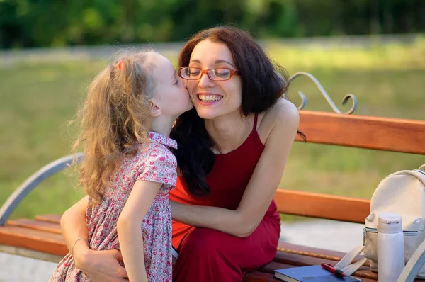 Retrato de la linda niña besando a su madre joven en gafas de vista y vestido rojo. Familia feliz sentada en el banco en el parque de la ciudad . — Foto de Stock