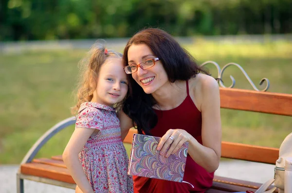 Retrato exterior de la joven madre y su linda hijita mirando a la cámara. La mujer sostiene un libro. Niño tiene dos colas de caballo . — Foto de Stock