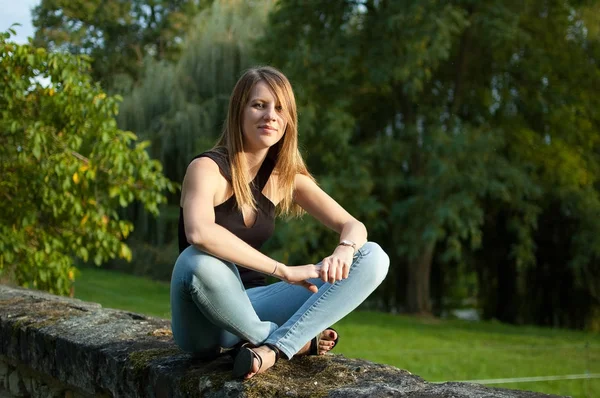 Retrato da jovem mulher bonito sentado no parque durante o pôr do sol em jeans e camisa preta . — Fotografia de Stock