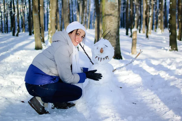 Усміхаючись молодої дівчини в білому капелюсі і шарф обіймає забавний сніговик в парку на сонячний день. Синє небо та морозний дерева на тлі. — стокове фото