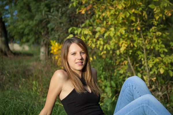 Draußen Porträt einer glücklichen schönen Frau in schwarzem Hemd und Jeans mit langen, glänzenden Haaren, die in der Nähe des Herbstbaums posiert und in die Kamera blickt. — Stockfoto