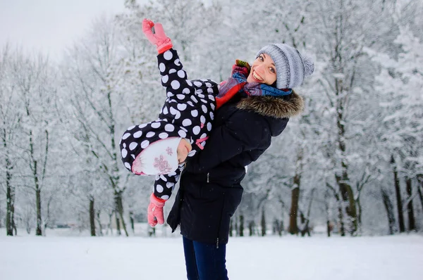 Mladá krásná matka hraje venku se svou malou dcerou v parku Snowy během dne. Aktivní maminka drží Funny Girl v rukou a usmívá se zubatými úsměv. — Stock fotografie