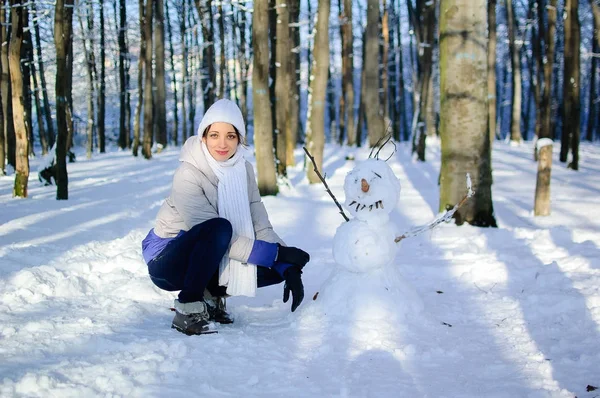 Beyaz şapka ve atkı komik kardan adam yanında oturan dışında bir kız portresi. Mavi gökyüzü ve arka plan ayaz ağaç. — Stok fotoğraf