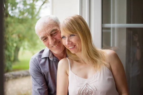 Pretty Beautiful Blonde De pie cerca de la ventana con su marido mayor y sonriendo. Concepto de diferencia de edad . —  Fotos de Stock