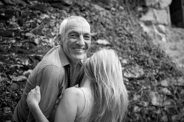 Retrato en blanco y negro del apuesto hombre mayor abrazando a su joven novia al aire libre y sonriendo mirando a la cámara. Pareja feliz con diferencia de edad . — Foto de Stock