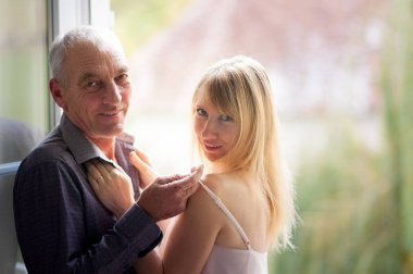 Portrait of Elderly Man Standing near the Window with His Young Blonde-haired Wife in Summer Short Dress. Couple with Age Difference. clipart
