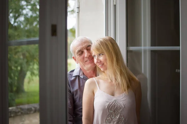 Счастливая пара с возрастом, обнимающаяся, стоя у открытых окон в комнате в летнее время. Концепция психологии отношений . — стоковое фото