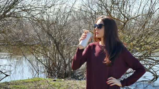 Jeune femme brune dans les lunettes de soleil boire une boisson en utilisant Thermo Cup sur fond de lac à l'extérieur. Concept de mobilité — Video