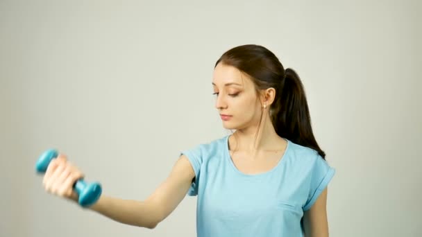 Молодая спортсменка с синим гантели в руке делает упражнения на сером фоне студии. Красотка с хвостиком в спортивной одежде. Концепция построения тела . — стоковое видео