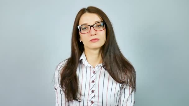 Серйозна молода дівчина в білій сорочці з червоним і синіми горизонтальними смугами дивлячись камера над її окулярів в студії на сірий фон. — стокове відео