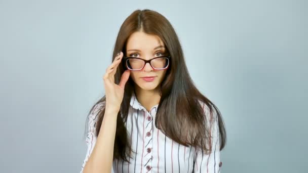 Attraente ragazza in camicia bianca con strisce rosse e blu guardando la fotocamera con interesse sopra i suoi occhiali con sorriso affascinante in studio su sfondo grigio . — Video Stock