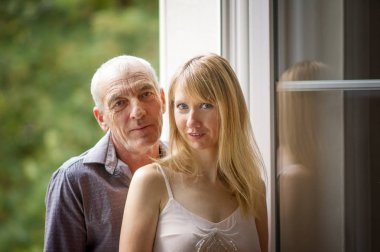 Portrait of Attractive Blonde Young Woman in Dress and Senior Man in Jeans and Blue Shirt Embracing near the Window. Age Difference Concept clipart