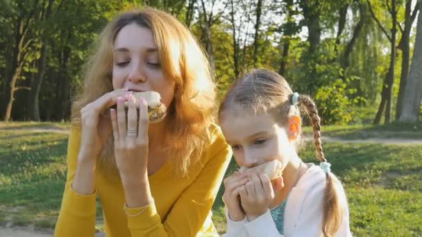 Närbild utomhus porträtt av ung blond kvinna äter hamburgare sitter på bänken i parken med sin lilla dotter. Ohälsosamt ätande, lunchkoncept — Stockvideo