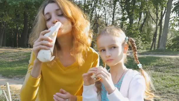 Närbild utomhus porträtt av ung blond kvinna äter shawarma sitter på bänken i parken med sin lilla dotter. Ohälsosamt ätande, lunchkoncept — Stockvideo