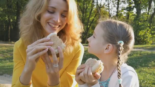 Szczęśliwa młoda blondynka matka i jej córeczka jedzą fast food siedząc na ławce w parku jesienią lub wiosną. Niezdrowe odżywianie, koncepcje czasu lunchu — Wideo stockowe