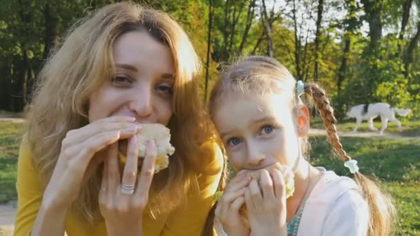 Gros plan portrait d'une heureuse jeune mère blonde et de sa petite fille mangeant à jeun assis sur le banc du parc à l'automne ou au printemps. Alimentation malsaine, concepts de l'heure du déjeuner 