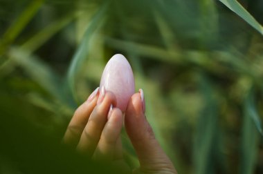 Vumfit, imbuilding veya meditasyon için pembe kuvars pembe yoni yumurta tutan Fransız manikür ile Kadın el. Yeşil ellerde Kristal mücevher açık havada arka plan kaynaklanıyor.