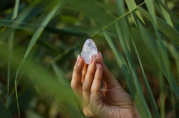 Mano femminile con manicure francese che tiene trasparente viola uovo ametista yoni per vumfit, imbuilding o meditazione. Uovo di quarzo cristallo in mano su sfondo verde all'aperto — Foto Stock