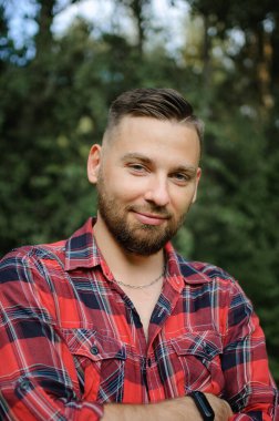 Kırmızı tişörtlü genç sakallı adamın portresini yakın çekimde ağaçların arkasındaki parktaki gülümsemeyle kameraya bakarak.