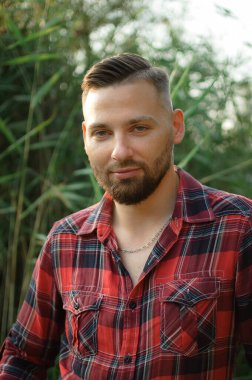 Kırmızı gömlekli vahşi sakallı genç adam yeşil saplı arka planda dışarıda duruyor. Hipster tarzı konsept.