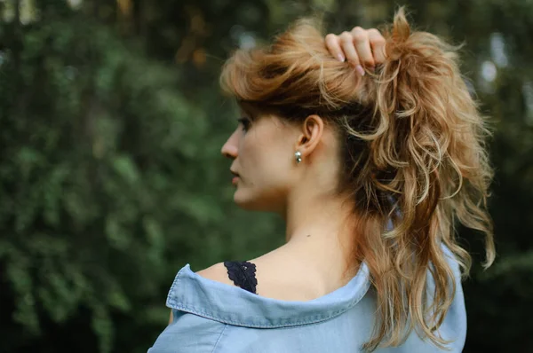 Side view portrait of sexy blonde girl with curly hair wearing blue casual jeans blouse and black lingerie posing in the park on green trees background. Hipster style concept. — Stock Photo, Image