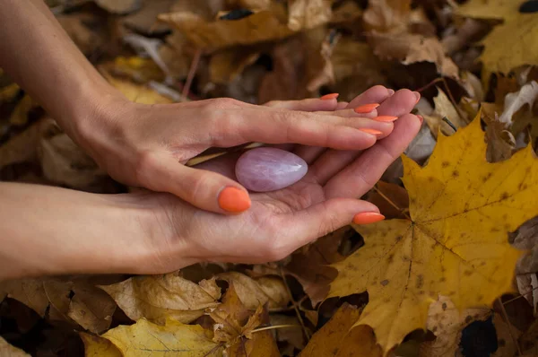 Female hand with orange manicure holding pink quartz yoni egg for vumfit, imbuilding or meditation on yellow fallen leaves background during autumn day outdoors