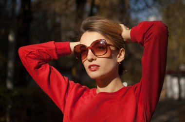 Havalı güneş gözlüklü çekici bir genç kadının açık hava kadın portresi. Çekici bir gülüşü, kısa sarı saçları, şehvetli dudakları ve sıcak güneşli bir günde sokakta poz veren kırmızı elbiseleri.