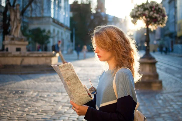 Вид сбоку молодой женщины в обычном свитере ищет направление с помощью бумажной карты ранним утром в древнем европейском городе на пустой площади — стоковое фото