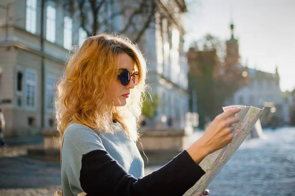 Menina adulta em azul sunglassed está olhando para o mapa de papel e à procura de direção no início da manhã na antiga cidade europeia na praça vazia — Fotografia de Stock