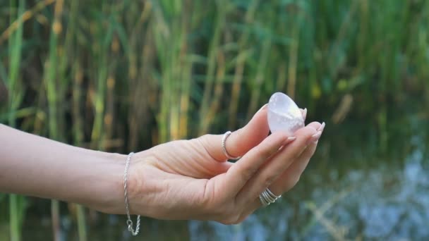 Mano femenina sosteniendo un huevo de cristal de cuarzo rosa yoni sobre el fondo del río. Salud de las mujeres, unidad con los conceptos de la naturaleza — Vídeos de Stock