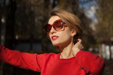 Havalı güneş gözlüklü çekici bir genç kadının açık hava kadın portresi. Çekici bir gülüşü, kısa sarı saçları, şehvetli dudakları ve sıcak güneşli bir günde sokakta poz veren kırmızı elbiseleri.