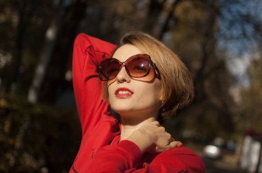 Havalı güneş gözlüklü çekici bir genç kadının açık hava kadın portresi. Çekici bir gülüşü, kısa sarı saçları, şehvetli dudakları ve sıcak güneşli bir günde sokakta poz veren kırmızı elbiseleri.