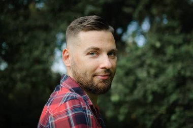 Kırmızı gömlekli vahşi sakallı genç adam dışarıda yeşil ağaçların arkasında duruyor. Hipster tarzı konsept.