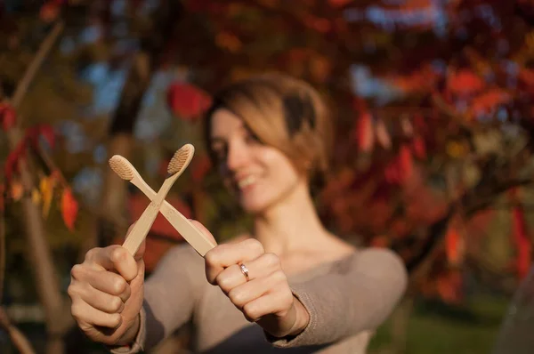 Young girl with short blond hair is holding a bamboo toothbrushes outdoors during sunny weather in autumn on red and orange leaves background. Environmental friendliness and zero waste concept