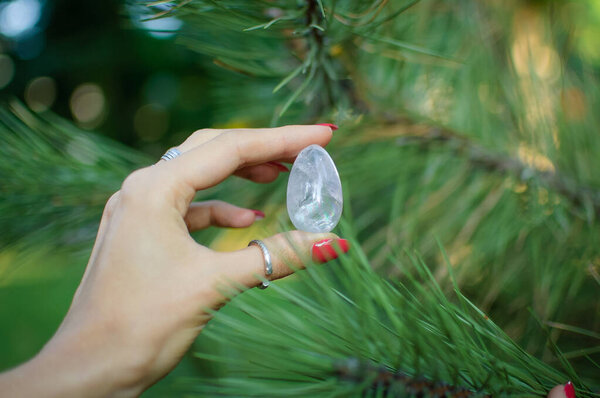 Female hand with transparent amethyst quartz yoni egg for vumfit, imbuilding or meditation. Shining crystal egg in hands on pine tree branches background outdoors. Womens health concept