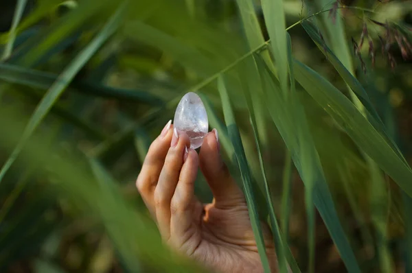 Mano femminile con manicure francese che tiene trasparente viola uovo ametista yoni per vumfit, imbuilding o meditazione. Uovo di quarzo cristallo in mano su sfondo verde all'aperto — Foto Stock