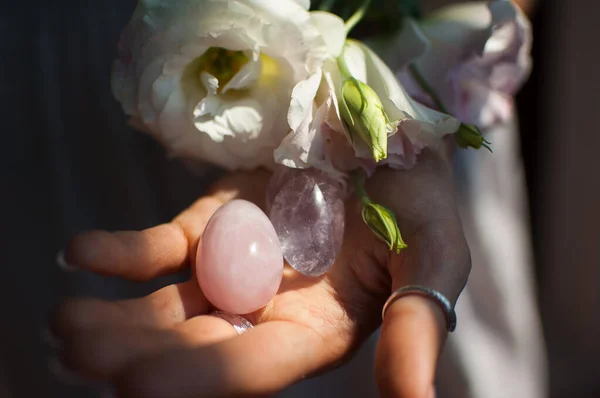 Mãos femininas segurando dois ovos yoni para vumfit, imbuilding ou meditação são feitas de quartzo rosa e ametista violeta transparente com flores brancas dentro de casa — Fotografia de Stock