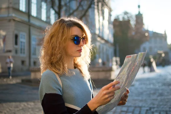 Menina adulta em azul sunglassed está olhando para o mapa de papel e à procura de direção no início da manhã na antiga cidade europeia na praça vazia — Fotografia de Stock