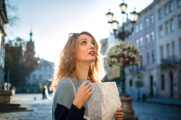 Menina adulta feliz animado está olhando para o mapa de papel e à procura de direção no início da manhã na antiga cidade europeia na praça vazia — Fotografia de Stock