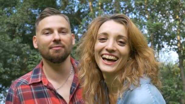 Charmant couple de petit ami hipster barbu en chemise rouge et petite amie blonde vêtue d'un jean décontracté bleu regardant la caméra sur fond d'arbres verts — Video